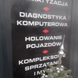 Czarna tablica informacyjna z białymi napisami: Klimatyzacja, Diagnostyka komputerowa, Holowanie pojazdów, Kompleksowe sprzątanie i mycie pojazdów, udekorowana małą choinką z srebrnymi bombkami.