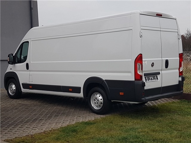 Biały samochód dostawczy typu furgon, zaparkowany na kostce brukowej z widocznym fragmentem trawnika, sfotografowany z perspektywy tylno-bocznej.