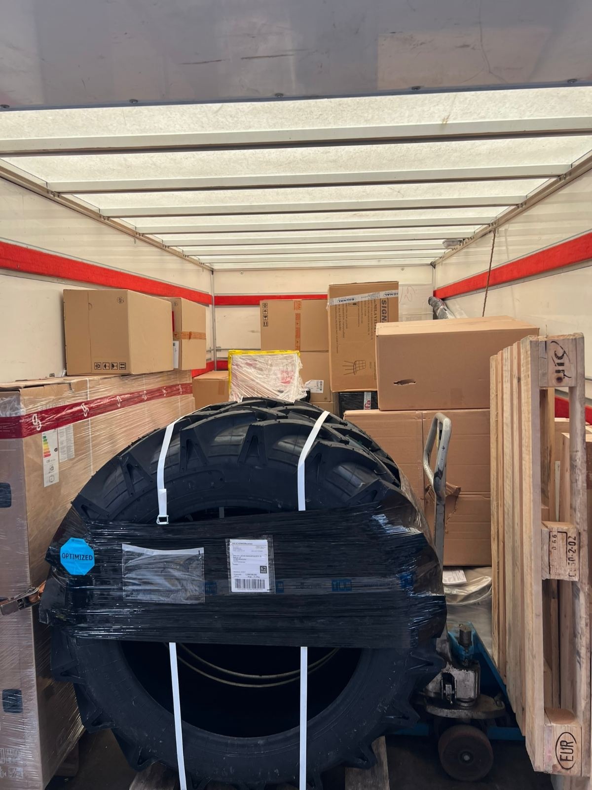 Wnętrze przestrzeni ładunkowej wypełnione kartonami i owiniętym folią zestawem opon rolniczych, zabezpieczonych pasami transportowymi, na palecie obok wózka transportowego.