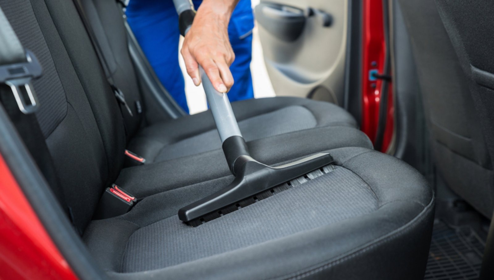 Czyszczenie tapicerki samochodowej odkurzaczem z nasadką szczotkową, widoczne tylne siedzenie, pasy bezpieczeństwa oraz fragment czerwonych drzwi.