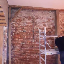 Odsłonięta ceglana ściana z drewnianą konstrukcją wzmacniającą, widoczny fragment rusztowania i osoba w czarnym ubraniu w trakcie prac remontowych.