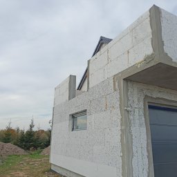 Alka -bud - Surowy stan budynku w trakcie budowy z białych bloczków, widoczne okno i brama garażowa, szare niebo w tle.