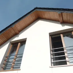 Fragment elewacji domu z dwoma oknami zabezpieczonymi metalowymi kratami, widoczna podbitka dachowa z oświetleniem punktowym i czarna dachówka, jasne tło nieba.