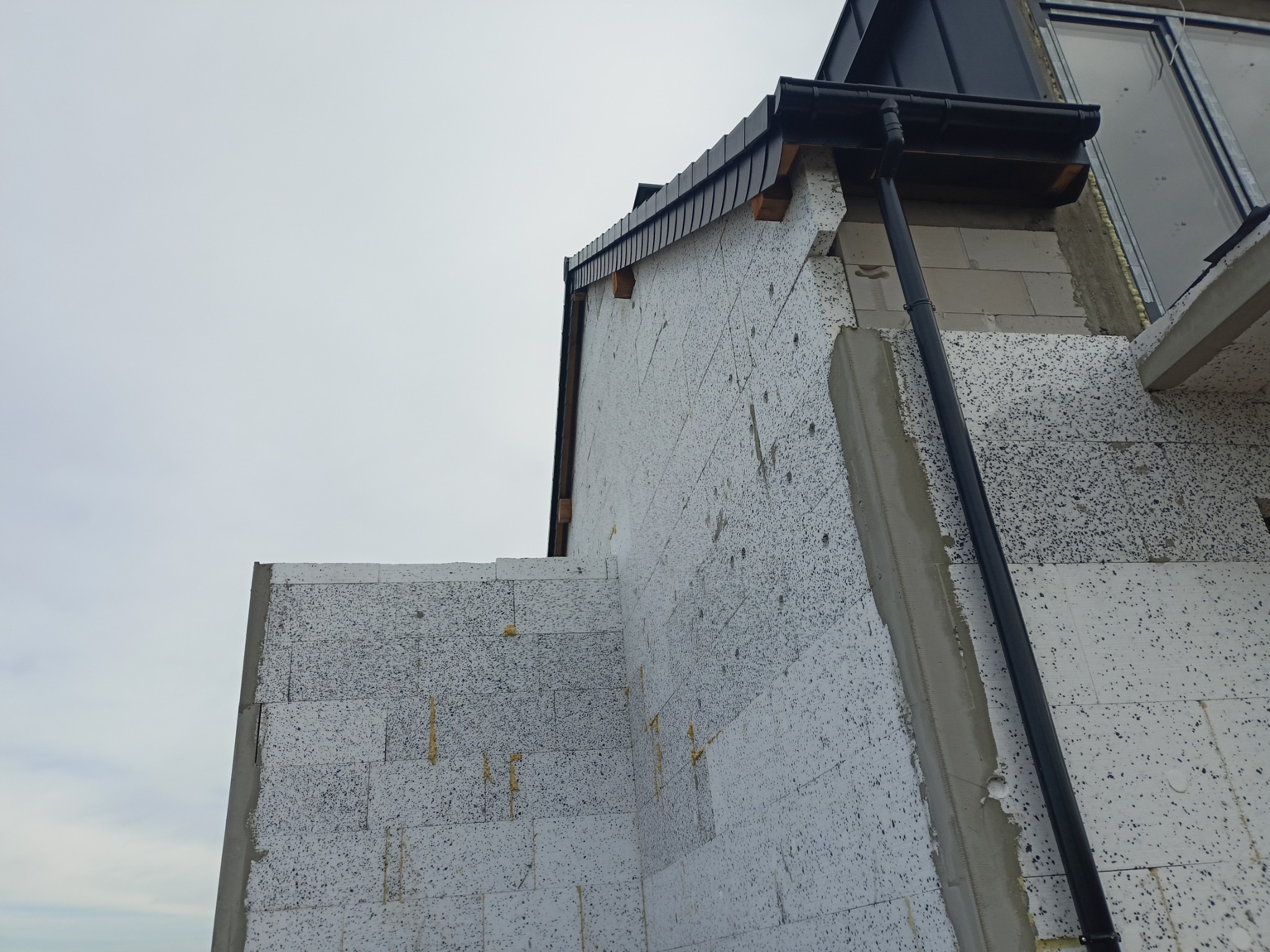 Niedokończona elewacja budynku z widocznym ociepleniem styropianowym, fragmentem orynnowania i surowym wykończeniem okiennym.