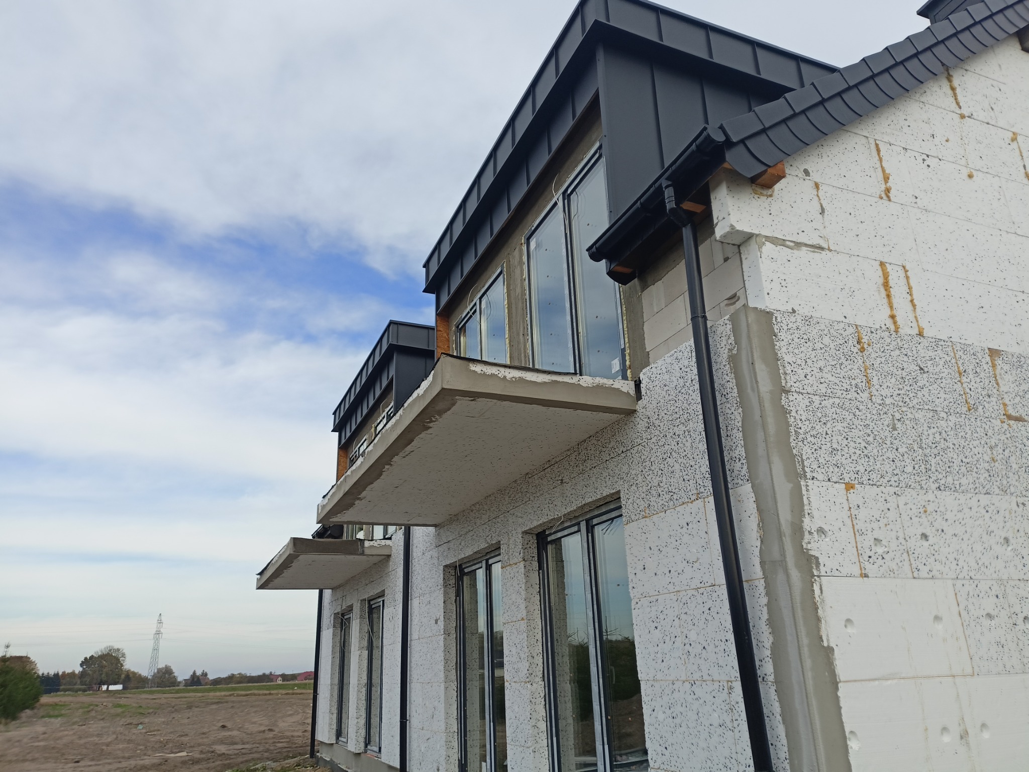 Surowy stan elewacji budynku z pustaków, widoczne okna z czarną stolarką, czarny dach i rynny, balkony w stanie deweloperskim, tło: krajobraz wiejski z liniami wysokiego napięcia.