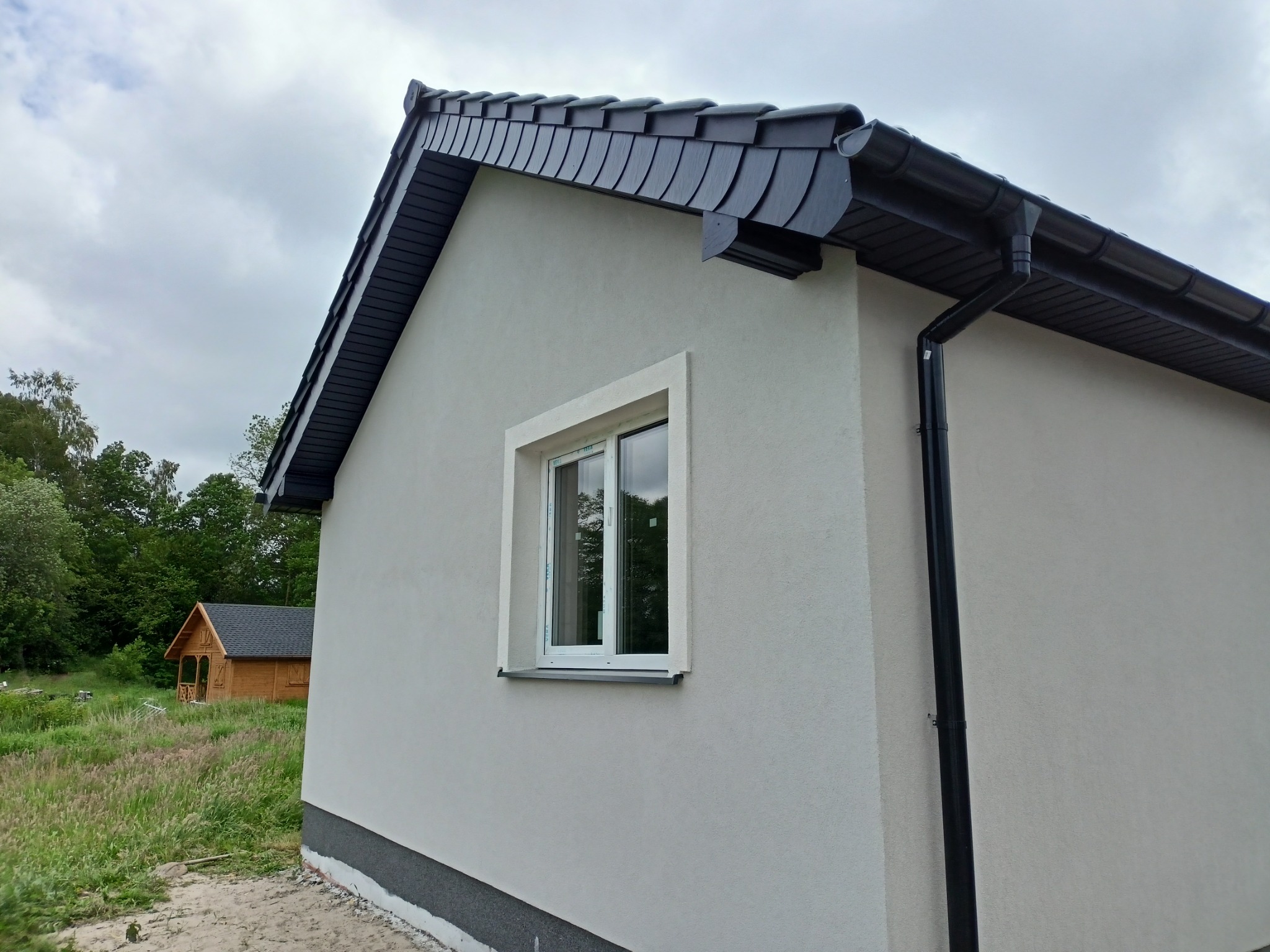 Nowo otynkowana ściana budynku z oknem w białej ramie, widoczny fragment dachu pokrytego czarną dachówką łuskową i czarna rynna spustowa, w tle drewniany domek.