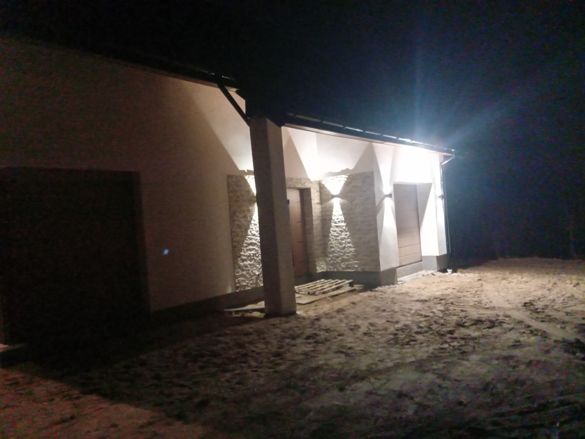 Nocny widok na elewację budynku z dekoracyjnym oświetleniem i fragmentami okładziny z kamienia dekoracyjnego. Widoczne dwa wejścia i teren wokół budynku.