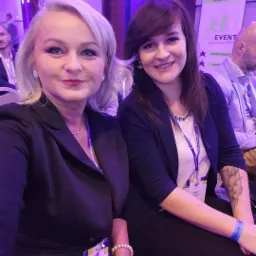 Dwie uśmiechnięte kobiety w strojach biznesowych, z identyfikatorami konferencyjnymi, siedzą na konferencji; widoczne logo organizatora w tle.