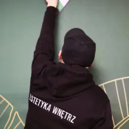 Osoba w czarnej bluzie z napisem 'ESTETYKA WNĘTRZ' wygładza zieloną ścianę narzędziem, widok z tyłu.