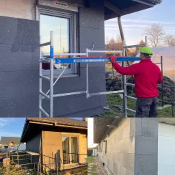 Pracownik w czerwonej bluzie i zielonej czapce montuje ciemnoszarą izolację termiczną na elewacji domu, stojąc na rusztowaniu aluminiowym.