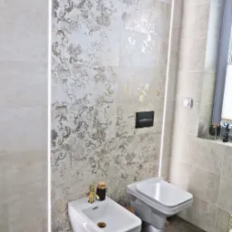 Nowoczesna łazienka z płytkami imitującymi stare złoto, podświetleniem LED, wiszącym bidetem z baterią w kolorze złota i toaletą.