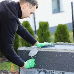 Pracownik w zielonych rękawicach nakłada zaprawę na szary blok izolacyjny, prawdopodobnie fundamentowy, używając szpachelki, w tle widoczne tuje i biały budynek.