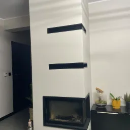 Nowoczesny kominek obudowany płytami w kolorze złamanej bieli z czarnymi akcentami, na tle czarnej szafki z roślinami i szklanym terrarium.