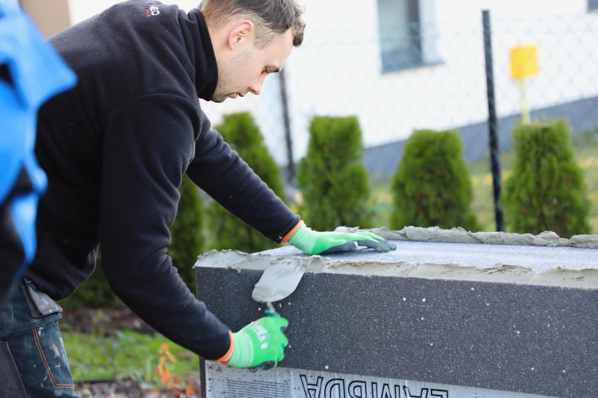 Pracownik w zielonych rękawicach nakłada zaprawę na szary blok izolacyjny, prawdopodobnie fundamentowy, używając szpachelki, w tle widoczne tuje i biały budynek.