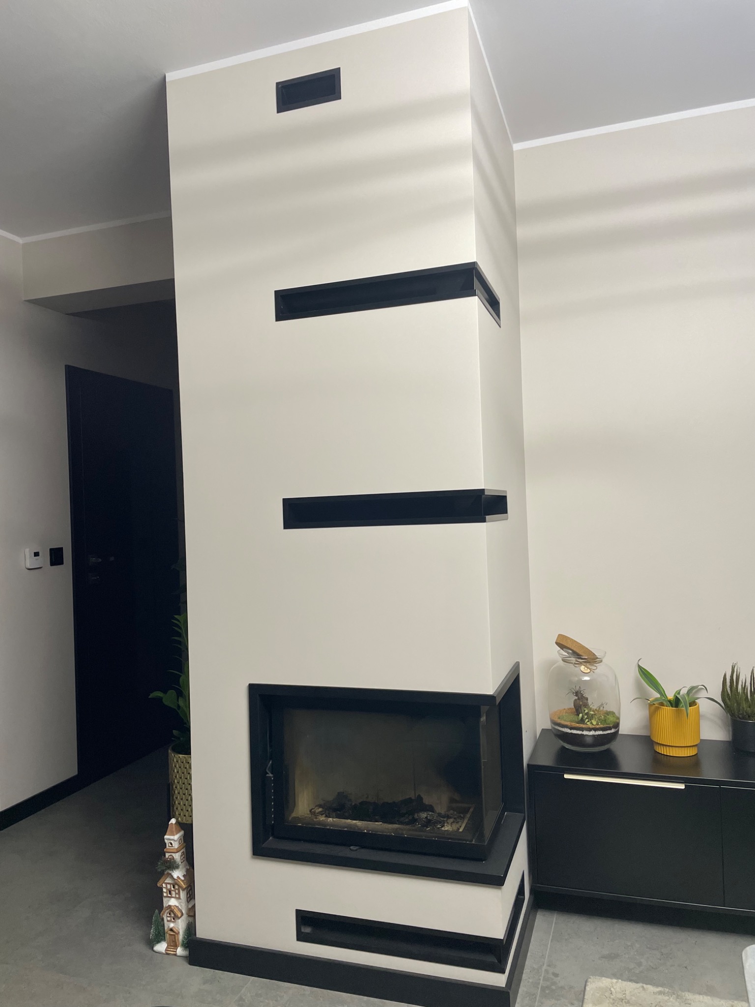 Nowoczesny kominek obudowany płytami w kolorze złamanej bieli z czarnymi akcentami, na tle czarnej szafki z roślinami i szklanym terrarium.