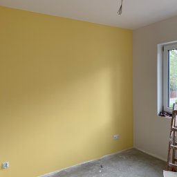 PAWEŁ NOWAKOWSKI USŁUGI REMONTOWE - Pokój po malowaniu: jedna ściana w odcieniu żółci, druga biała, widoczne gniazdka elektryczne, surowa podłoga, drabina malarska i wiadro z farbą w rogu.