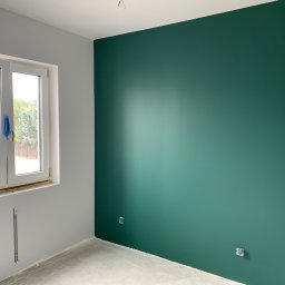 PAWEŁ NOWAKOWSKI USŁUGI REMONTOWE - Wnętrze pokoju po malowaniu: ściana w odcieniu butelkowej zieleni, szara ściana z oknem zabezpieczonym folią, surowa podłoga i wystające kable elektryczne.
