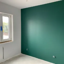 Wnętrze pokoju po malowaniu: ściana w odcieniu butelkowej zieleni, szara ściana z oknem zabezpieczonym folią, surowa podłoga i wystające kable elektryczne.