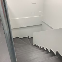 PAWEŁ NOWAKOWSKI USŁUGI REMONTOWE - Nowoczesne schody wykończone szarą płytką, białe ściany z minimalistycznymi panelami i metalowa poręcz.