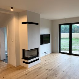 PAWEŁ NOWAKOWSKI USŁUGI REMONTOWE - Wnętrze salonu po remoncie: jasna podłoga z desek, białe ściany, nowoczesny kominek z czarnymi akcentami, duże okno balkonowe z widokiem na zewnątrz, wiszący telewizor i minimalistyczne oświetlenie...