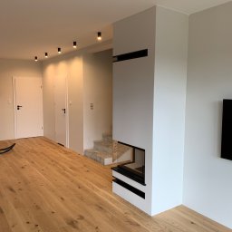 PAWEŁ NOWAKOWSKI USŁUGI REMONTOWE - Jasne wnętrze z drewnianą podłogą, białymi ścianami, nowoczesnym kominkiem i schodami w trakcie wykończenia.