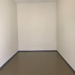 Pomalowane na szaro wnętrze pomieszczenia z taśmą malarską przy podłodze, jasne ściany i sufit z lampą