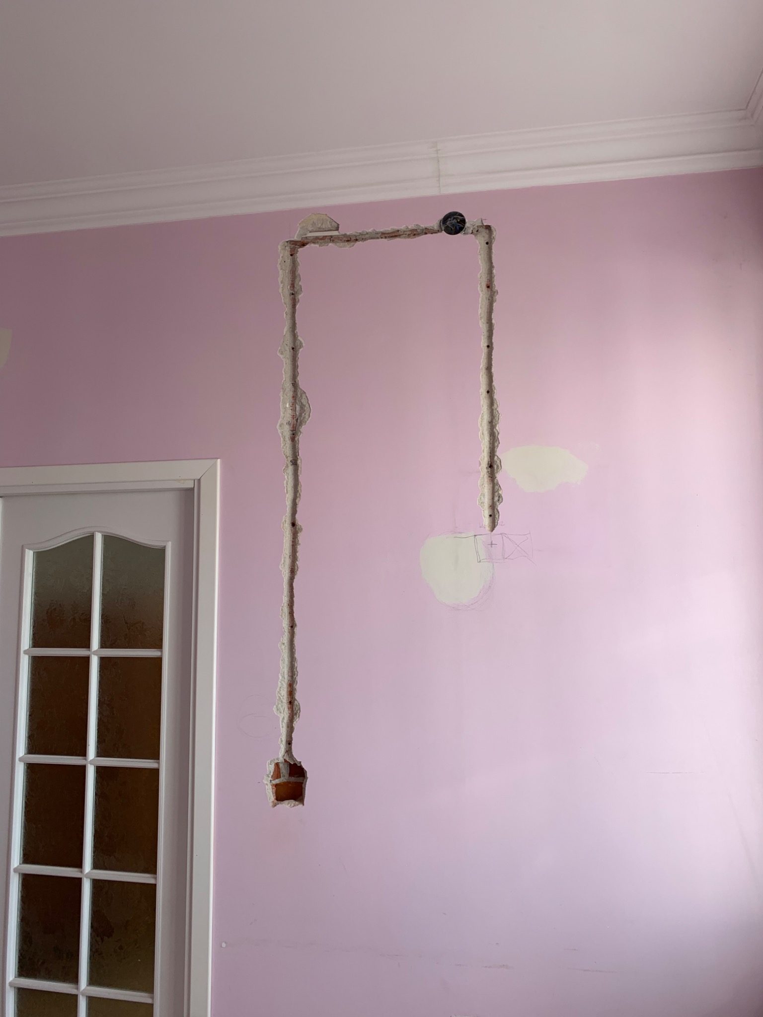 Różowa ściana z bruzdami po instalacjach elektrycznych przygotowana do wyrównywania, widoczne ślady po gipsie i rysunki pomocnicze pod gniazdko, obok białe drzwi.