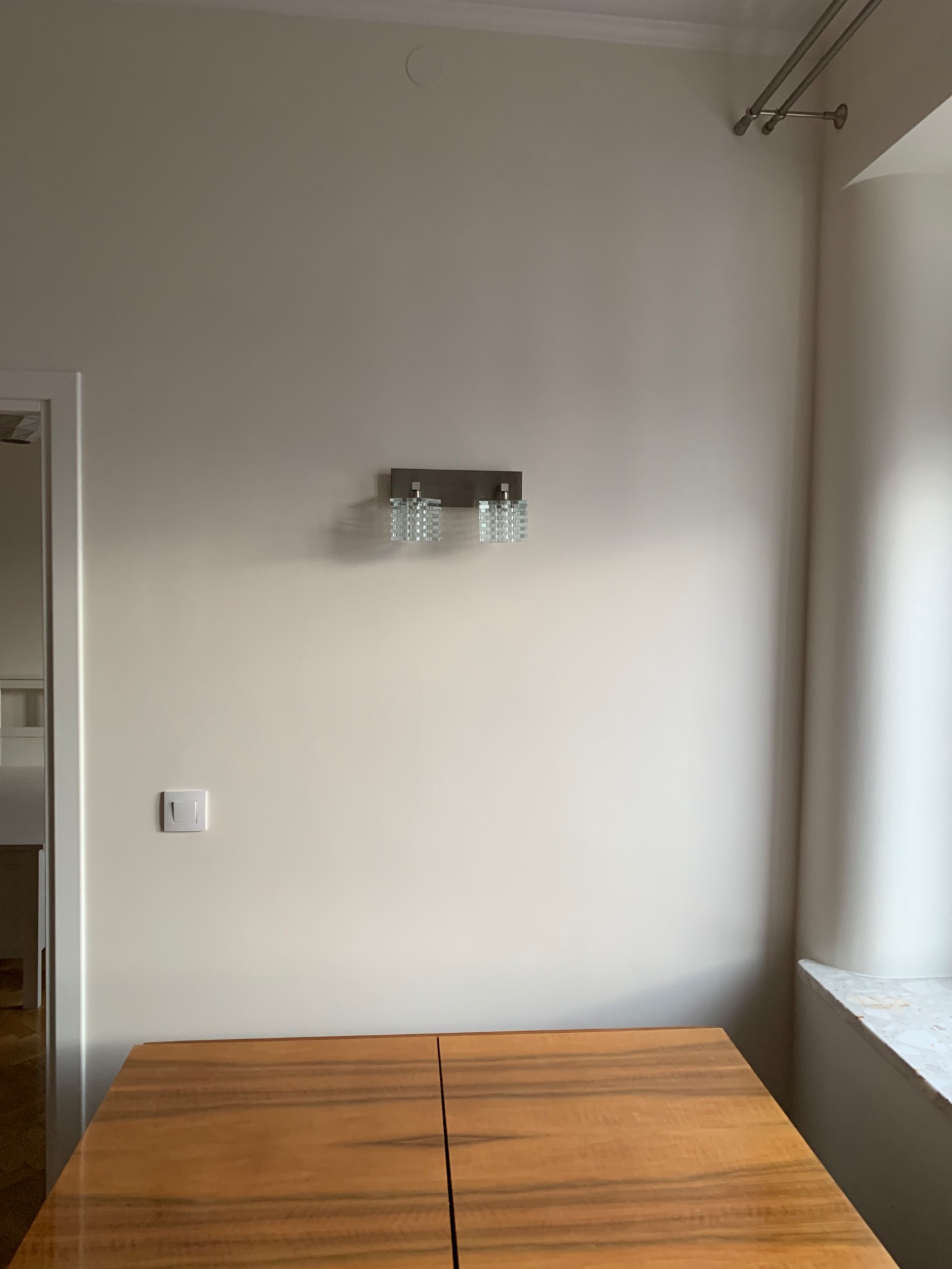 Jasna ściana z kremową farbą, na której wisi nowoczesny, kryształowy kinkiet, poniżej widoczny włącznik światła i fragment blatu drewnianego stołu, w tle widoczne okno z marmurowym parapetem.