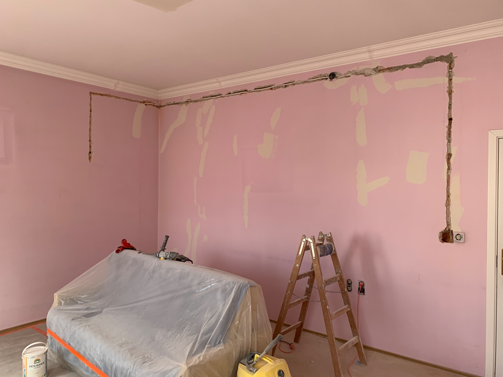 Różowa ściana w trakcie przygotowania do prac wykończeniowych, widoczne wyżłobione bruzdy pod instalacje elektryczne, częściowo wypełnione gipsem, zabezpieczona folią kanapa, drabina i narzędzia.