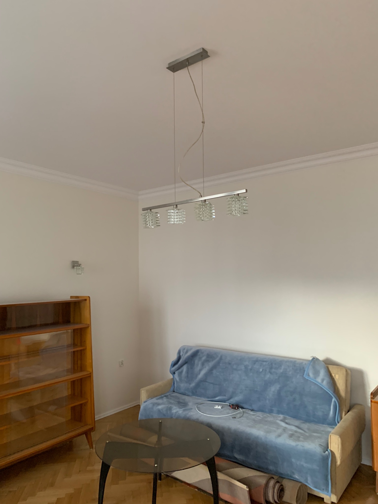 Pokój z jasnymi ścianami, drewnianą podłogą w jodełkę i modernistycznym oświetleniem sufitowym. Sofa przykryta niebieskim pledem, obok stolik kawowy i regał.