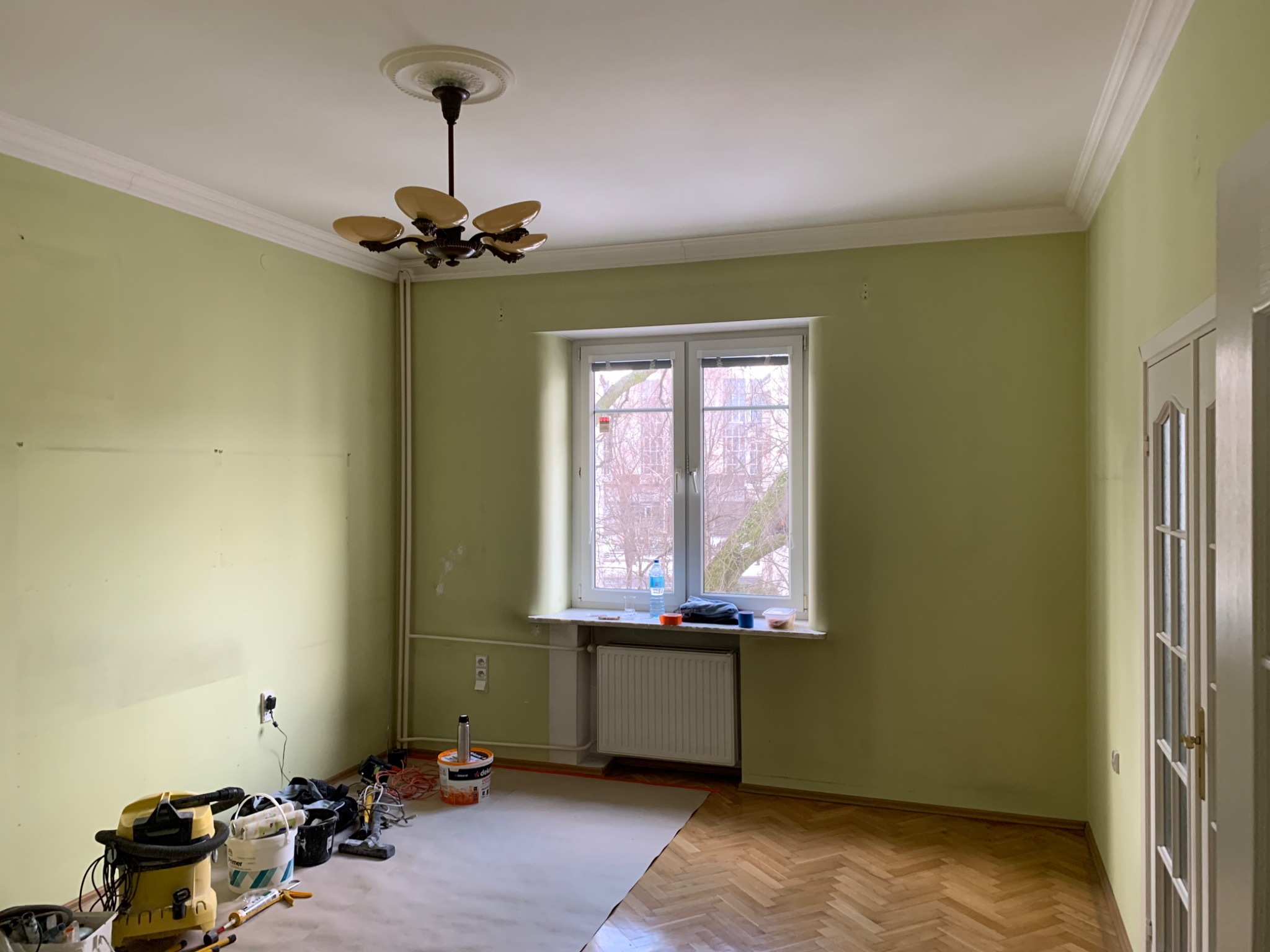 Pokój przed odświeżeniem: zielone ściany z drobnymi ubytkami, parkiet zabezpieczony kartonem, widoczne narzędzia i akcesoria malarskie, w tle okno z widokiem na drzewa.