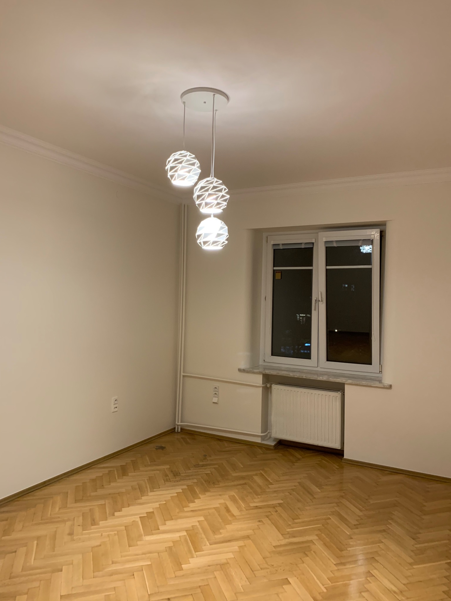 Pusty pokój z parkietem w jodełkę, świeżo pomalowanymi ścianami na biało i nowoczesną lampą wiszącą z trzema kulami oświetleniowymi.