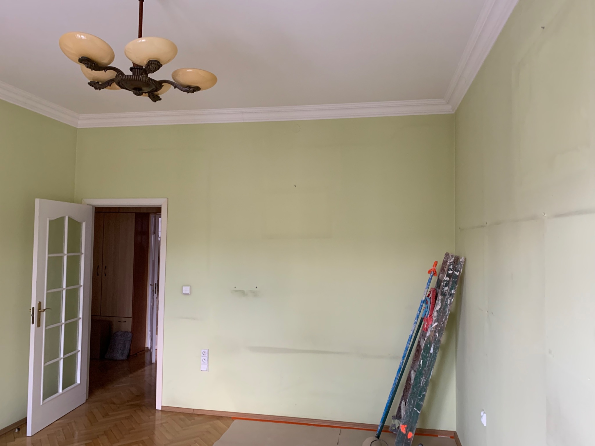 Pokój z zielonymi ścianami w trakcie przygotowania do malowania, widoczne ślady po wcześniejszych pracach, drabina oparta o ścianę, zabezpieczona podłoga i stara lampa sufitowa.