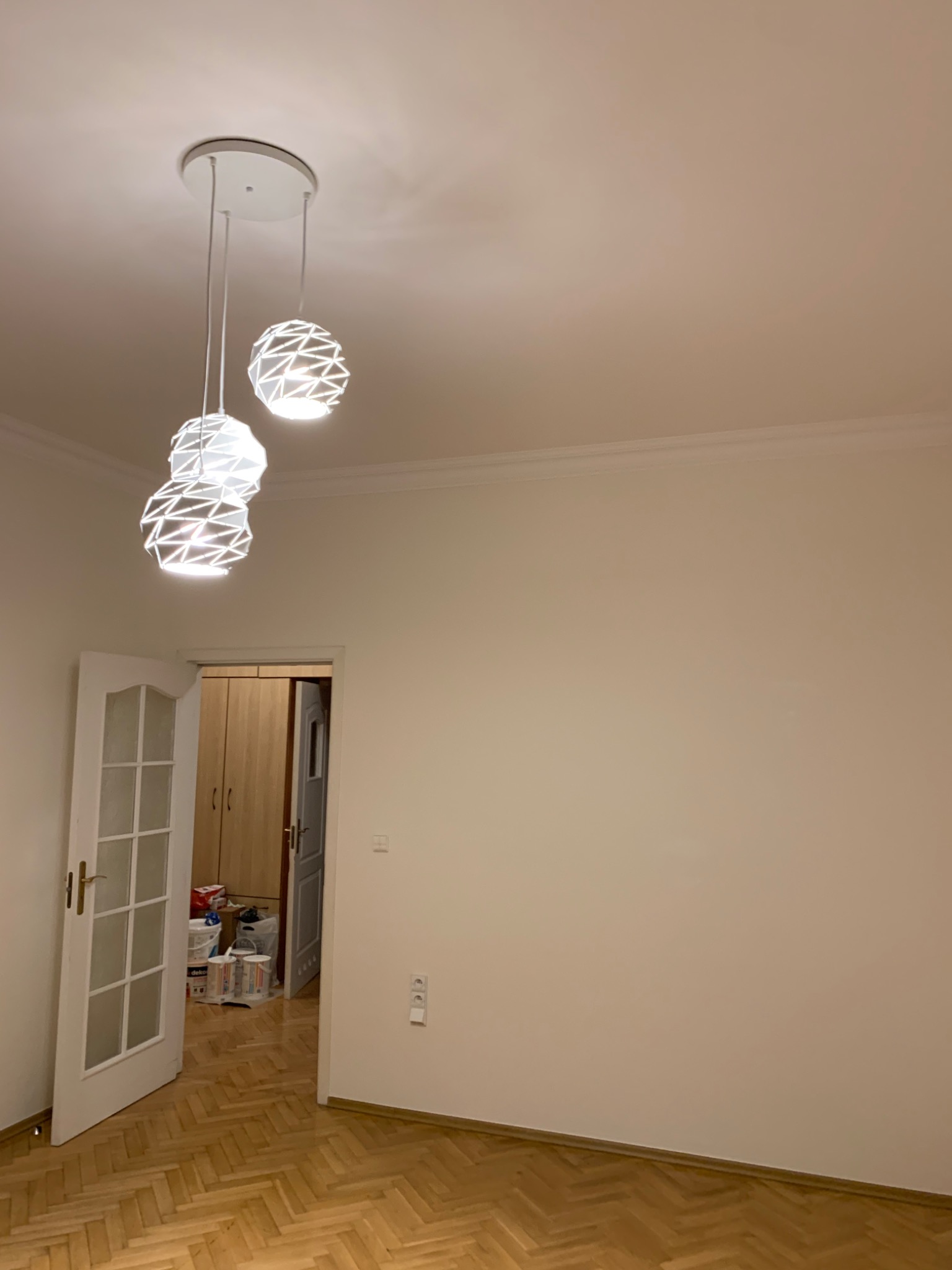 Pusty pokój z parkietem, świeżo pomalowanymi ścianami w odcieniu beżu i nowoczesną, potrójną lampą wiszącą z ażurowymi kloszami, widoczne otwarte drzwi do kolejnego pomieszczenia z wiadrami farby.