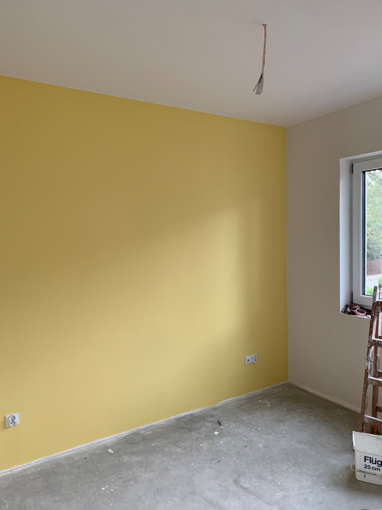 Pokój po malowaniu: jedna ściana w odcieniu żółci, druga biała, widoczne gniazdka elektryczne, surowa podłoga, drabina malarska i wiadro z farbą w rogu.