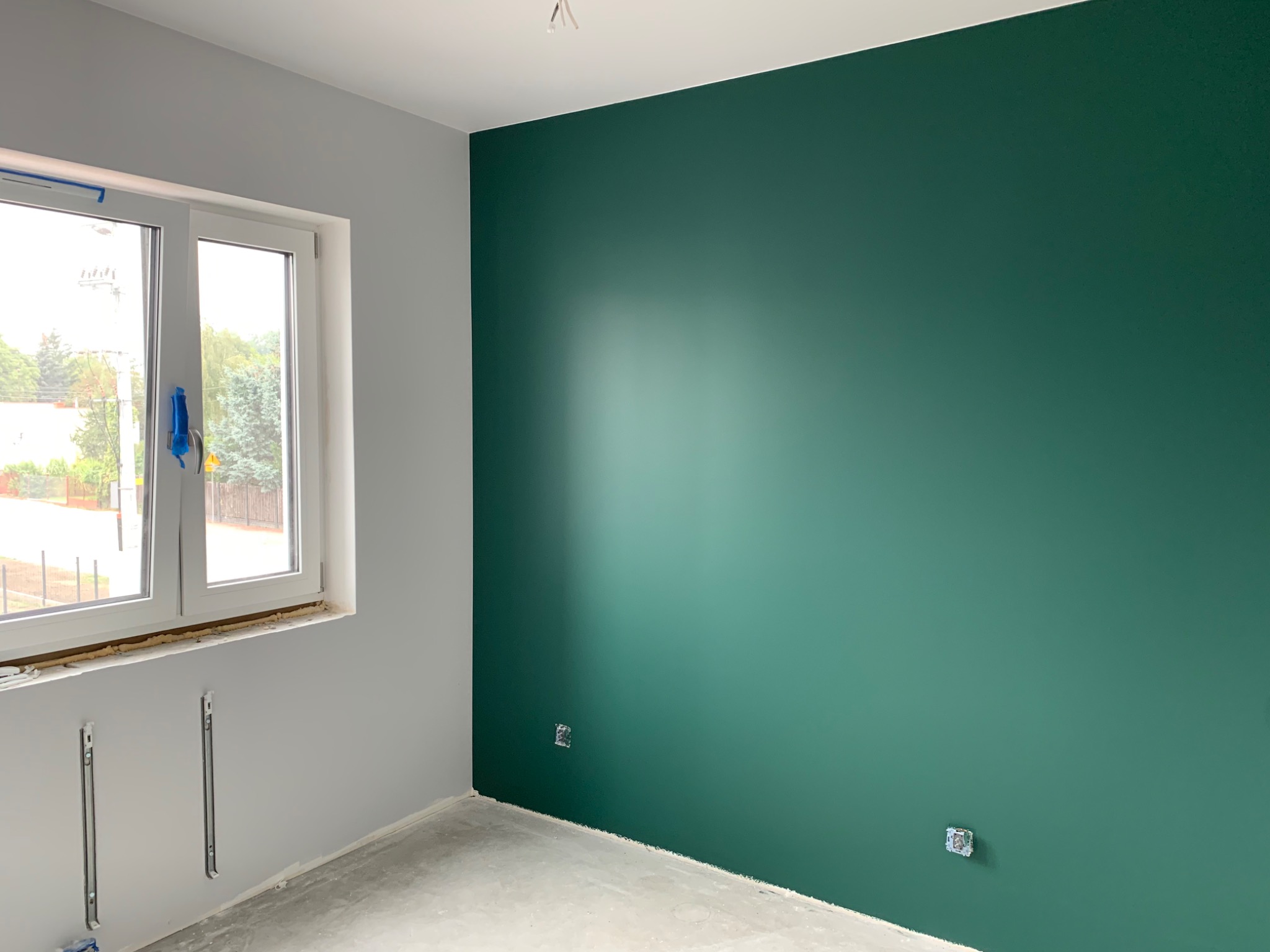 Wnętrze pokoju po malowaniu: ściana w odcieniu butelkowej zieleni, szara ściana z oknem zabezpieczonym folią, surowa podłoga i wystające kable elektryczne.