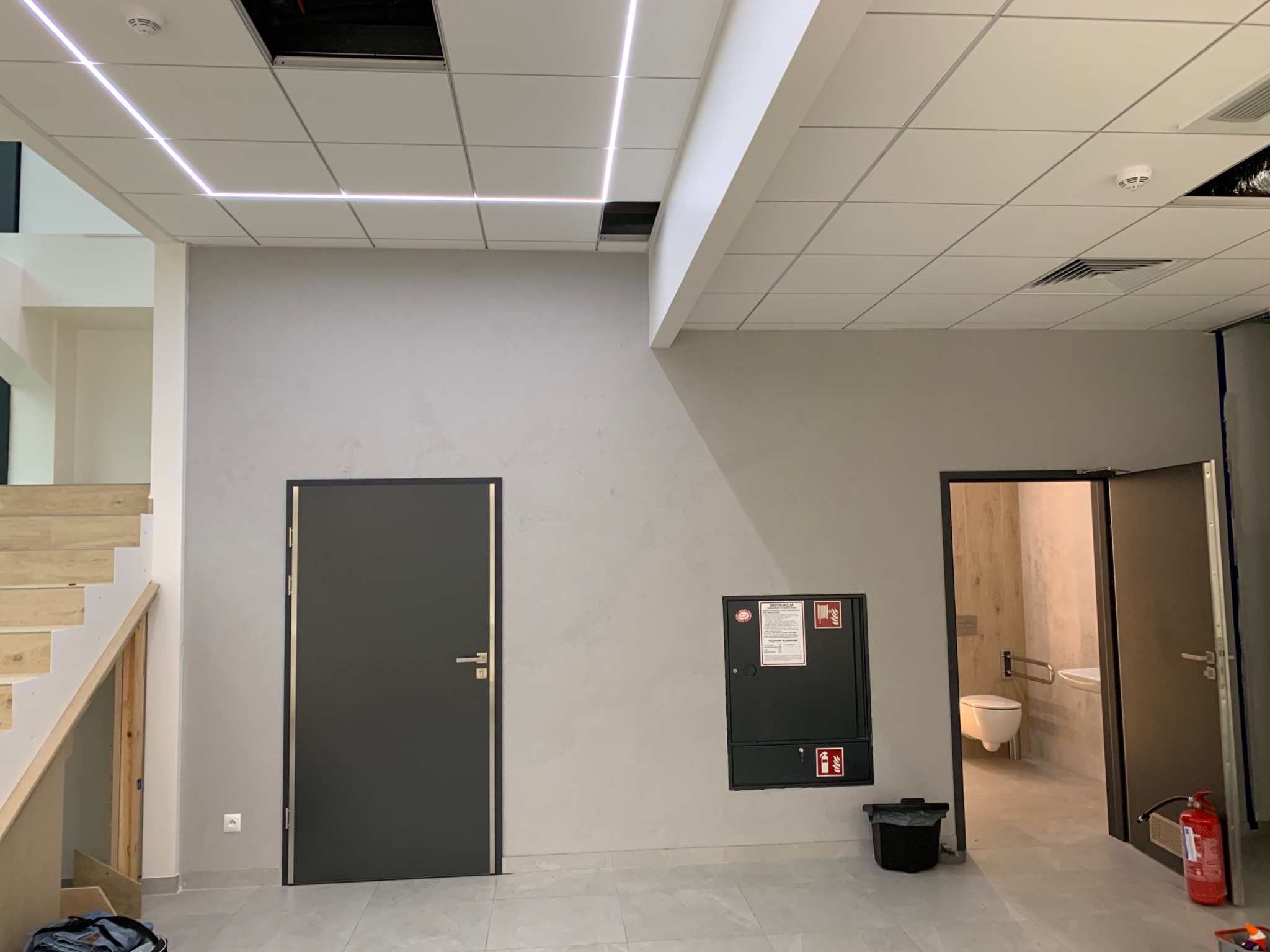 Wnętrze biurowe po odświeżeniu: gładka, szara ściana z dwiema ciemnymi drzwiami i szafką przeciwpożarową, podwieszany sufit z liniowym oświetleniem LED, widoczne schody drewniane i toaleta w tle.