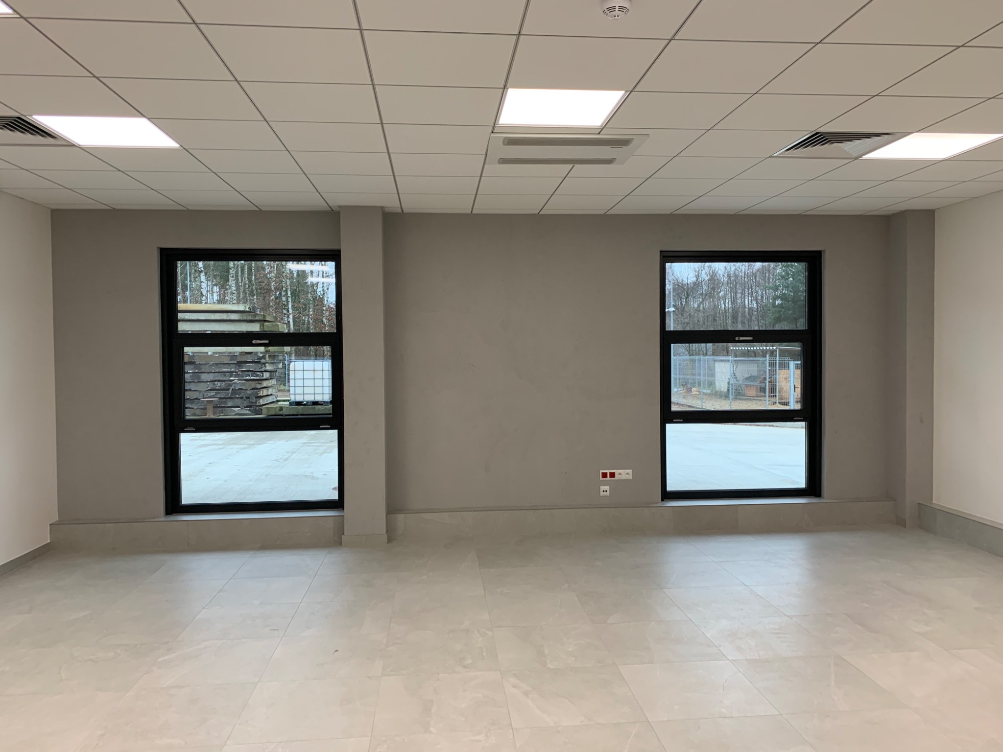 Puste, jasne pomieszczenie po remoncie z dwoma oknami w czarnych ramach, szarą ścianą z gładzią, podłogą wyłożoną płytkami ceramicznymi w odcieniach beżu i białym sufitem kasetonowym z oświetleniem...