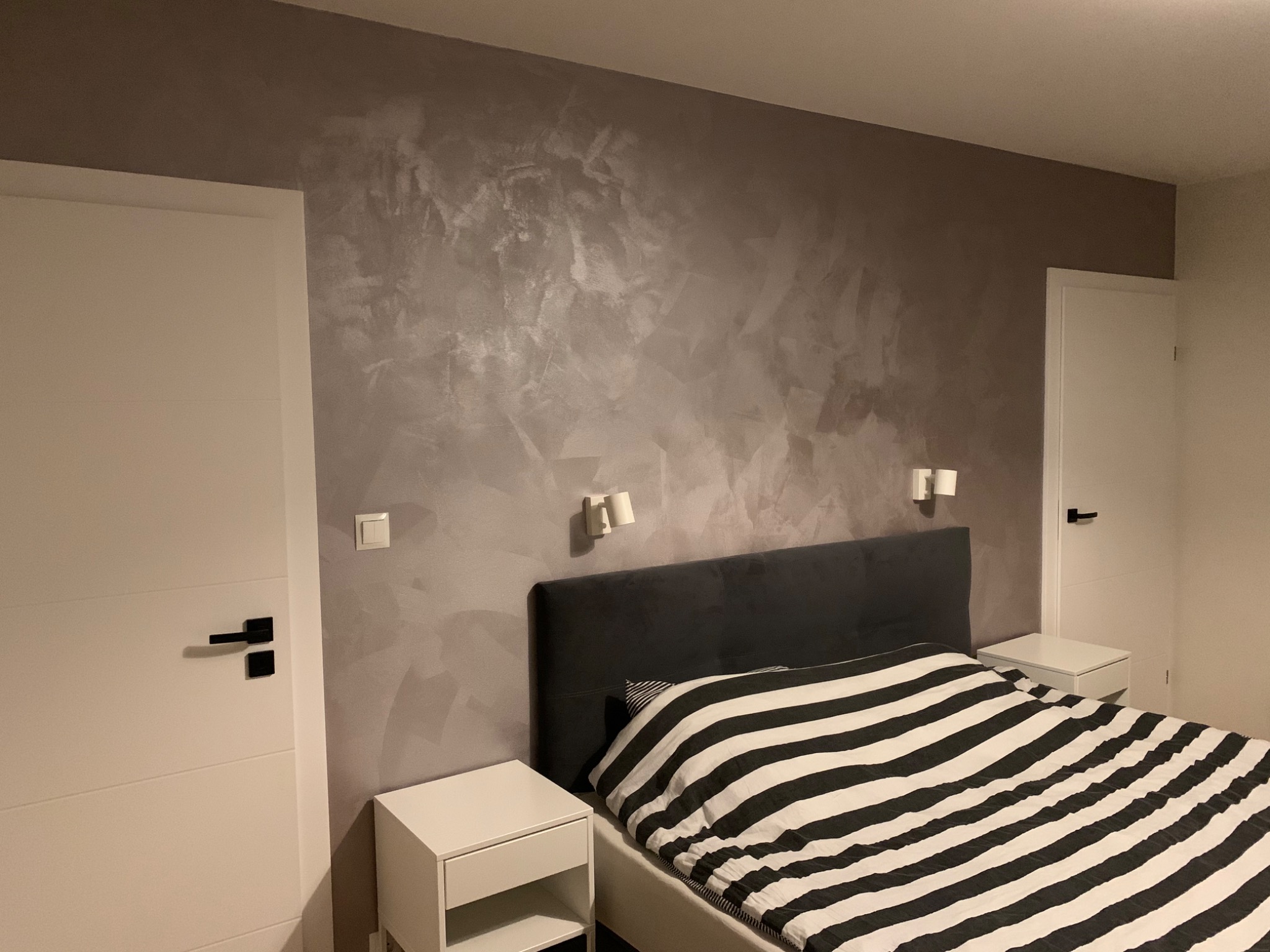 Sypialnia z dekoracyjną, szarą ścianą z efektem stiuku weneckiego, oświetlona kinkietami, z białymi drzwiami i minimalistycznymi szafkami nocnymi.