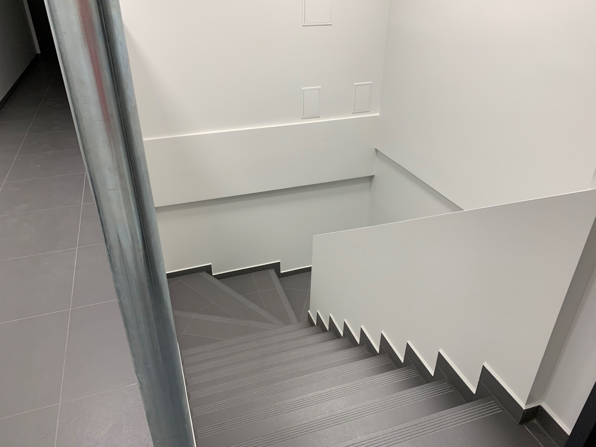 Nowoczesne schody wykończone szarą płytką, białe ściany z minimalistycznymi panelami i metalowa poręcz.