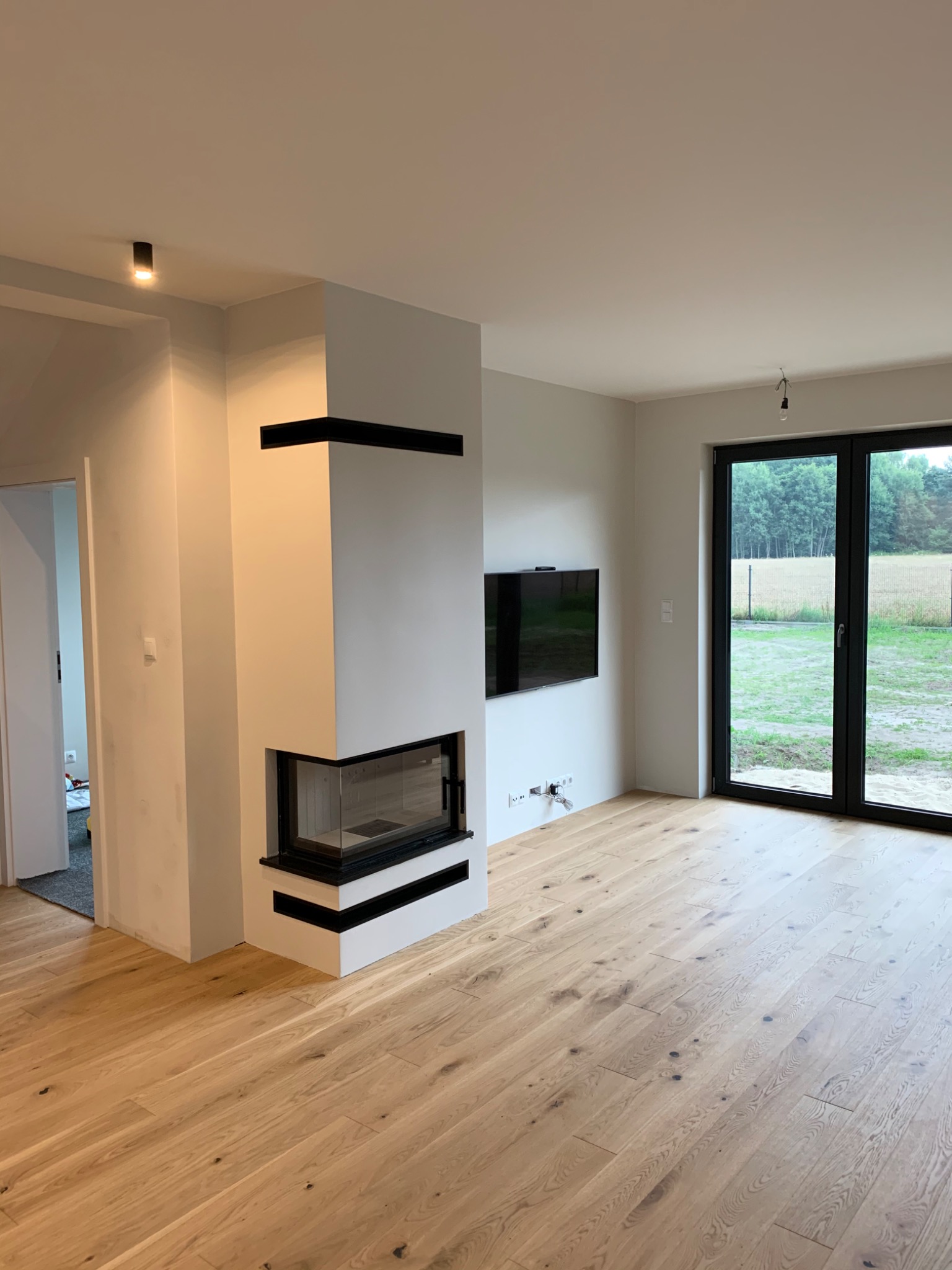 Wnętrze salonu po remoncie: jasna podłoga z desek, białe ściany, nowoczesny kominek z czarnymi akcentami, duże okno balkonowe z widokiem na zewnątrz, wiszący telewizor i minimalistyczne oświetlenie...