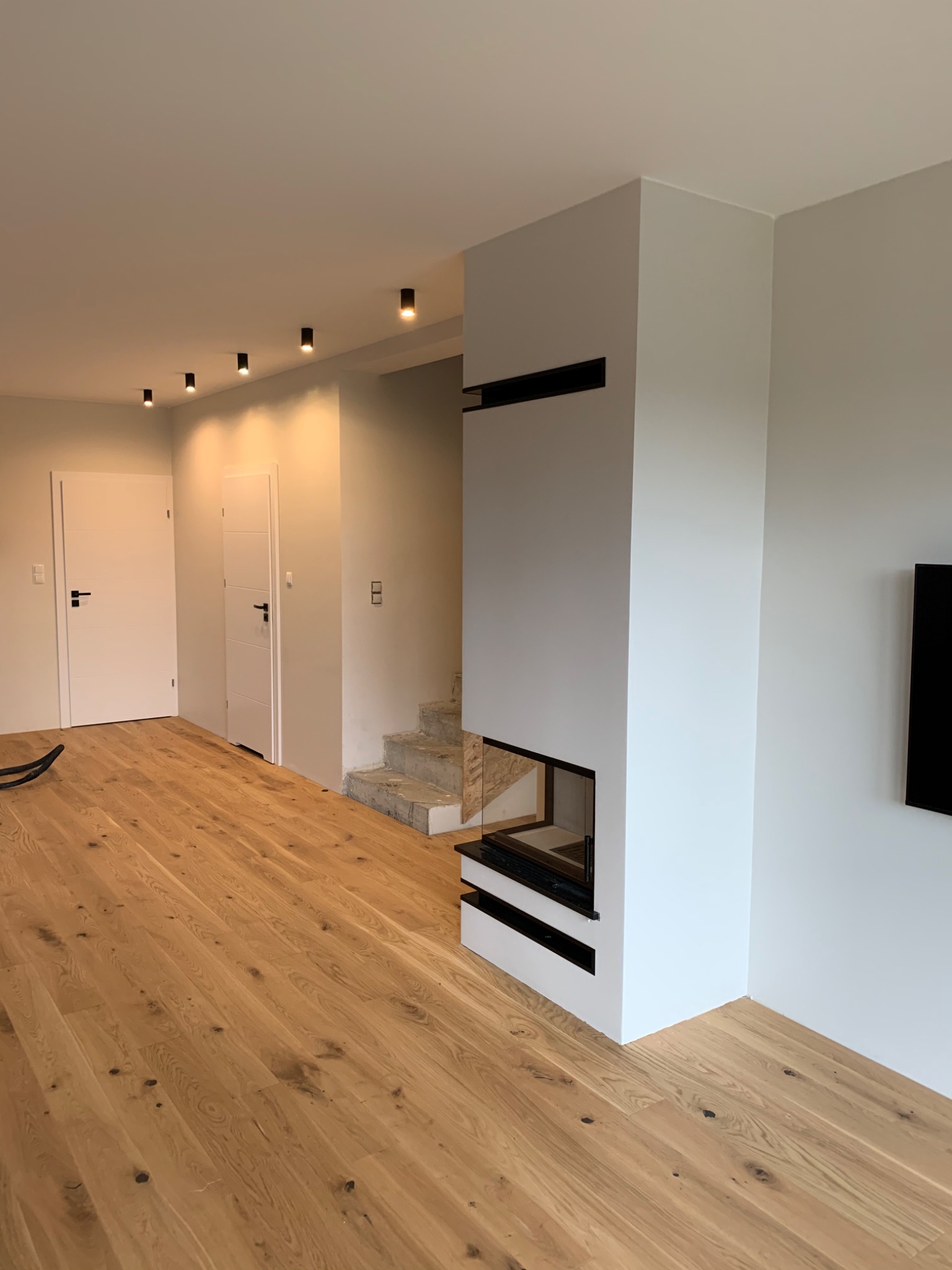 Jasne wnętrze z drewnianą podłogą, białymi ścianami, nowoczesnym kominkiem i schodami w trakcie wykończenia.