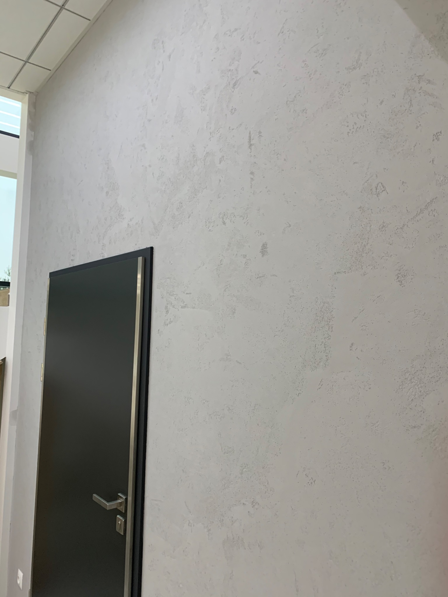 Jasnoszara ściana z dekoracyjną strukturą tynku mineralnego, obok czarne drzwi z metalowym obramowaniem i klamką.
