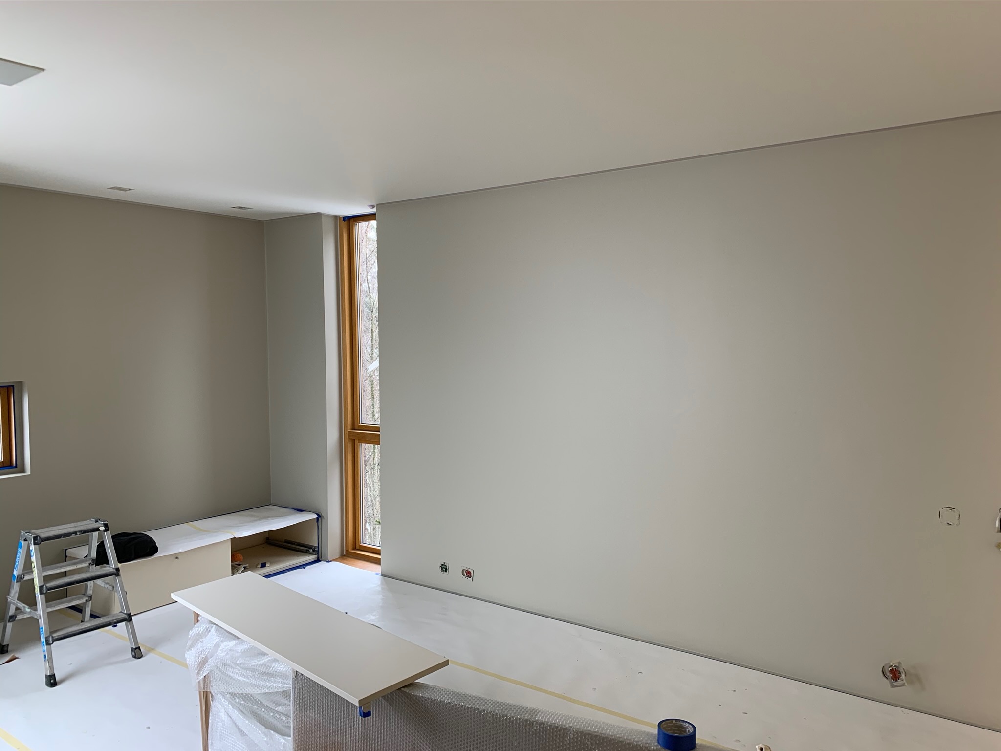 Przygotowane do malowania wnętrze w Kajetanach: zabezpieczona podłoga, drabina i blat roboczy przy ścianie z oknem, widoczne gniazdka elektryczne.