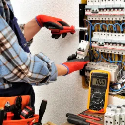 Elektryk w czerwonych rękawicach montuje bezpiecznik w skrzynce rozdzielczej, używając śrubokręta; obok leży pomarańczowy multimetr wskazujący zero.