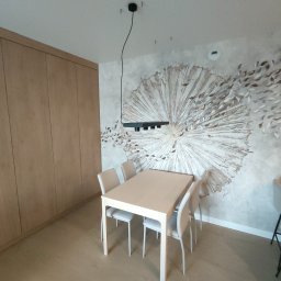 Jakub Sokół - Jasne, minimalistyczne wnętrze jadalni z drewnianą zabudową ścienną, stołem z krzesłami o jasnej kolorystyce oraz dekoracyjną tapetą z motywem rozchodzących się liści na ścianie. Nad stołem wisi...