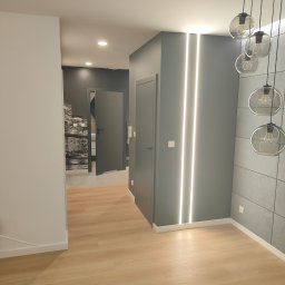 Jakub Sokół - Nowoczesny korytarz z panelami podłogowymi w odcieniach beżu, szarą ścianą z pionowym oświetleniem LED, wiszącą lampą z przezroczystymi kloszami i widokiem na pokój z fototapetą przedstawiającą...