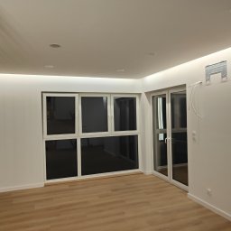 Jakub Sokół - Jasne wnętrze z panelami podłogowymi i dużymi oknami, widoczny uchwyt klimatyzacji na ścianie z wystającymi przewodami, oświetlenie ledowe pod sufitem.