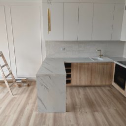 Jakub Sokół - Wnętrze jasnej kuchni w trakcie wykańczania, widoczna drabina oparta o ścianę z dekoracyjnymi listwami, marmurowy blat, drewniane szafki i panele podłogowe ułożone w jodełkę.