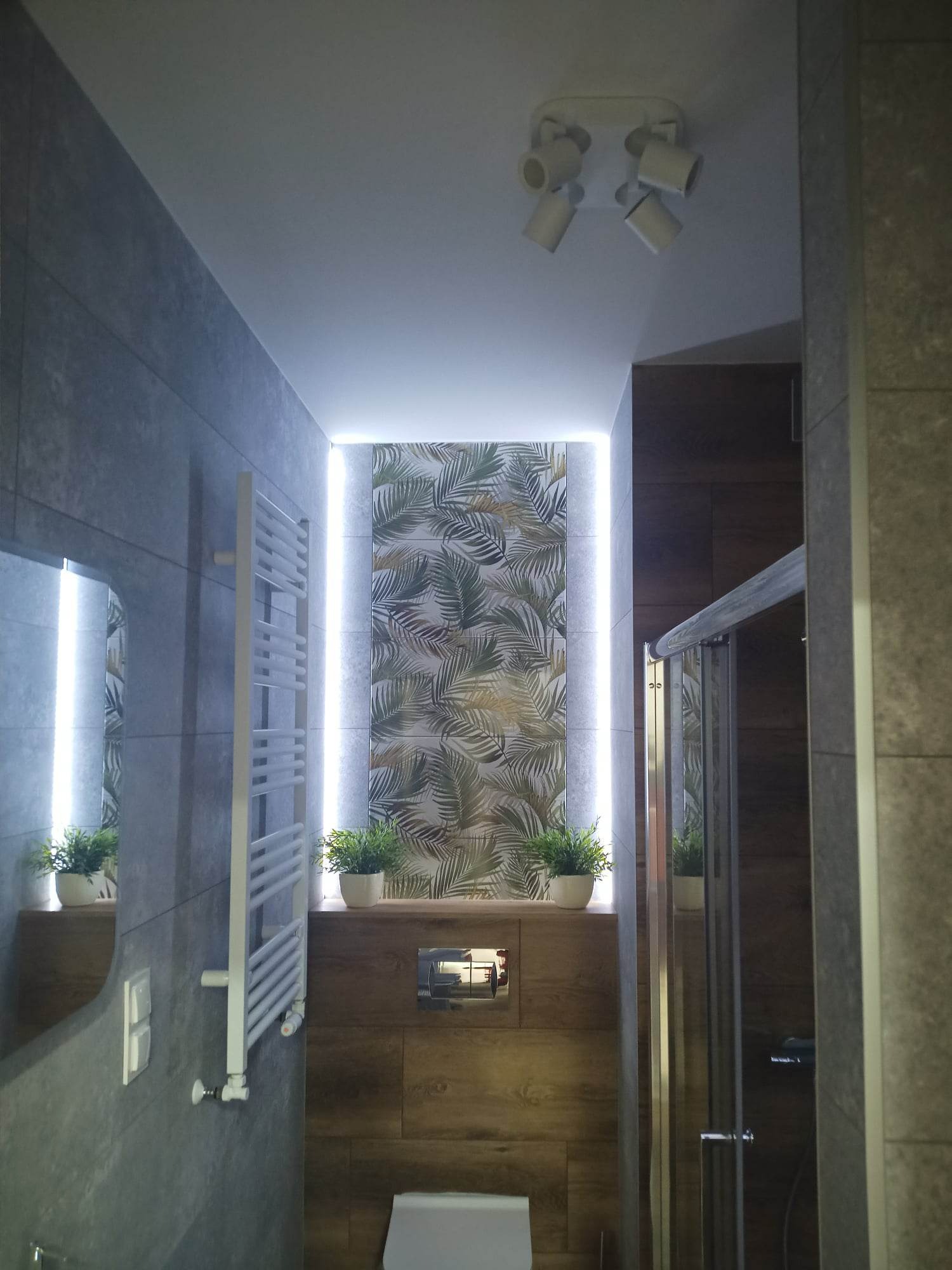 Nowoczesna łazienka z szarymi płytkami, panelem dekoracyjnym z motywem liści w podświetlanej wnęce, białym grzejnikiem drabinkowym i kabiną prysznicową z elementami szkła.